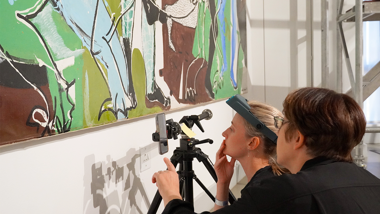 Two people looking closely at a the bottom of a painting while it hangs on the wall.