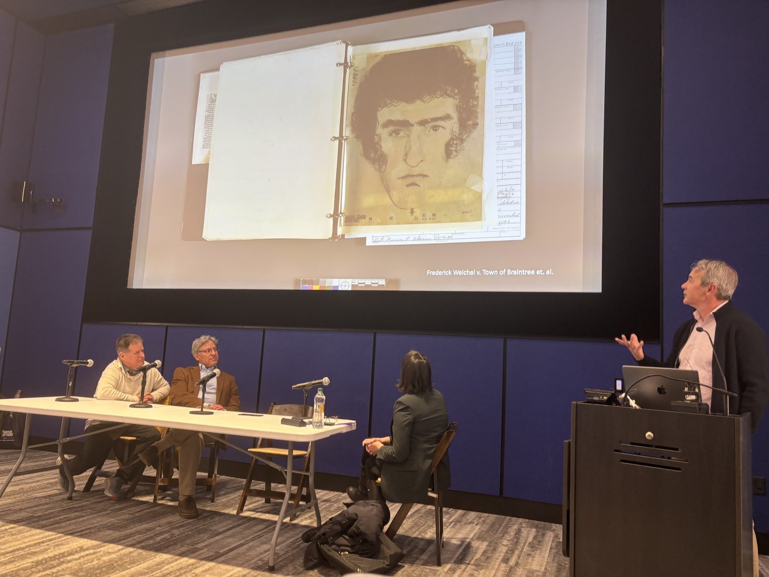 Panelists sit at a table looking at a projection while a speaker presents from a podium.