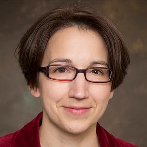 A woman with short brown hair and glasses wears a red coat while smiling at the camera.