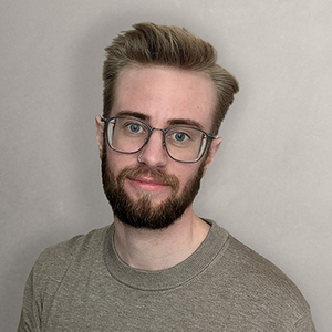 A man with facial hair, glasses, and a nose ring wears a gray short smiling directly at the camera. 