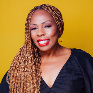 A woman with long light brown braids, red lipstick, and a black top smiles directly at the camera.