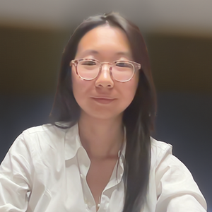 A woman with long dark brown hair, glasses, and a white collared shirt smiles directly at the camera.