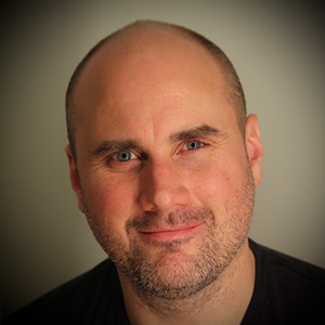 A man with short facial hair and a black shirt smiles directly at the camera.