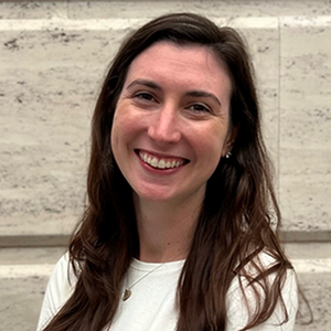 A woman with long brown hair and a white long sleeve shirt smiles directly at the camera.
