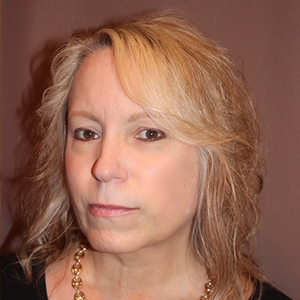 A woman with short blonde hair and a gold necklace looks seriously at the camera. 