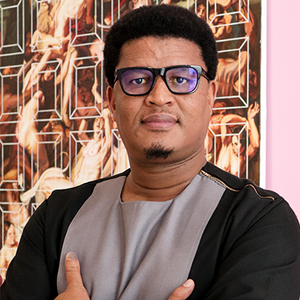 A man with dark skin and dark hair looks intensely with black glasses and a grey and black shirt.