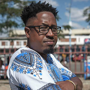 A man with dark skin and dark hair looks intensely over his shoulder with black glasses and a white and blue patterned shirt.