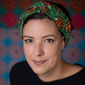 A woman with dark hair wears a colorful headband and black shirt while shirt smiling directly at the camera.
