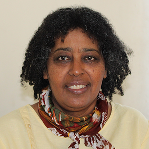 A woman with dark curly hair smiles at the camera. She wears a colorful scarf and yellow shirt.