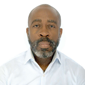 A bald man with dark skin and salt and pepper beard looks directly at the camera, wearing a white collared shirt.