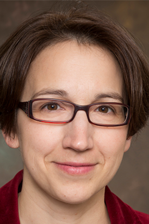 A woman with short brown hair and glasses wears a red coat while smiling at the camera.