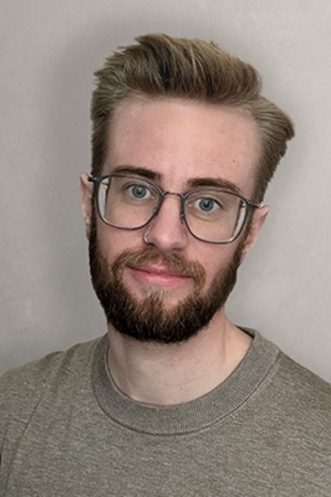 A man with facial hair, glasses, and a nose ring wears a gray short smiling directly at the camera. 