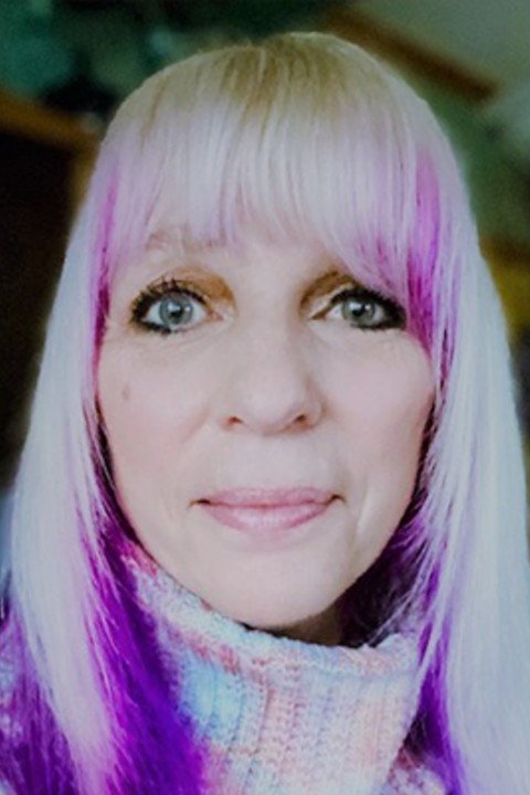 A woman with blond hair with pink and purple highlights and a turtleneck smiles directly at the camera.