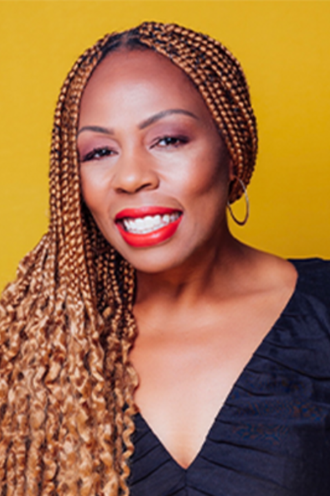 A woman with long light brown braids, red lipstick, and a black top smiles directly at the camera.