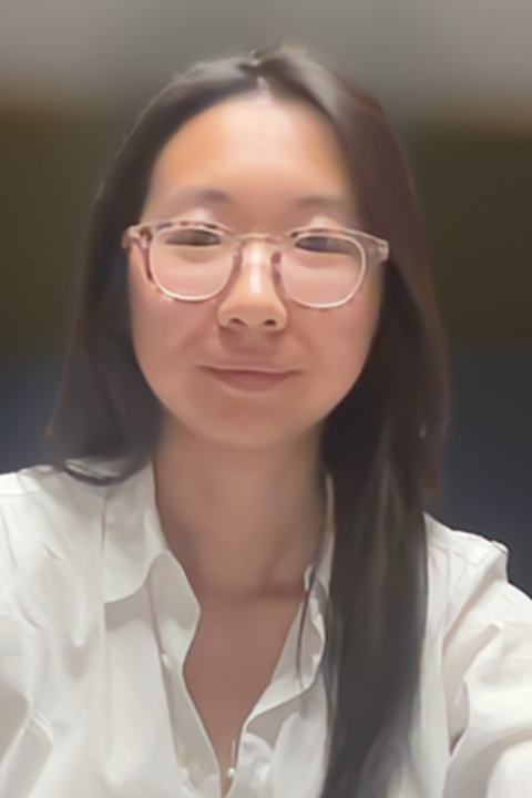 A woman with long dark brown hair, glasses, and a white collared shirt smiles directly at the camera.