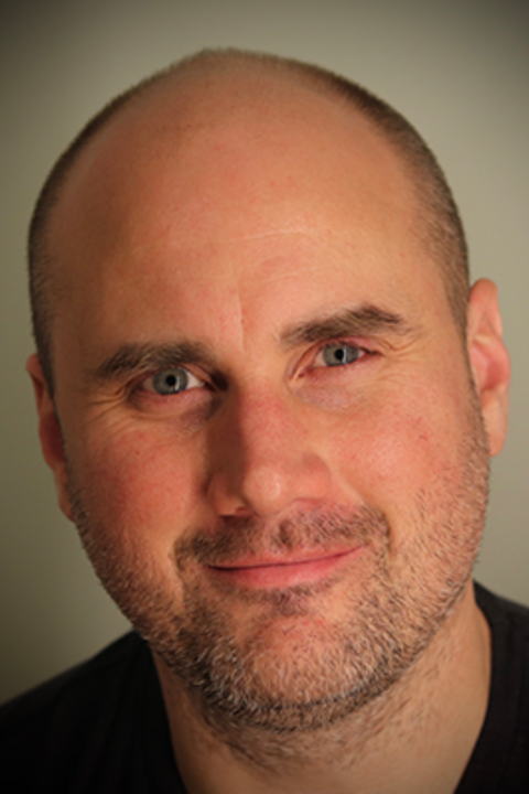 A man with short facial hair and a black shirt smiles directly at the camera.