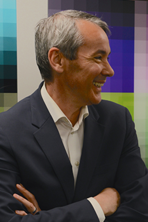 A man smiles over his left shoulder with short grey hair wears a dark blue shirt and white collared shirt in front of a colorful wall. 