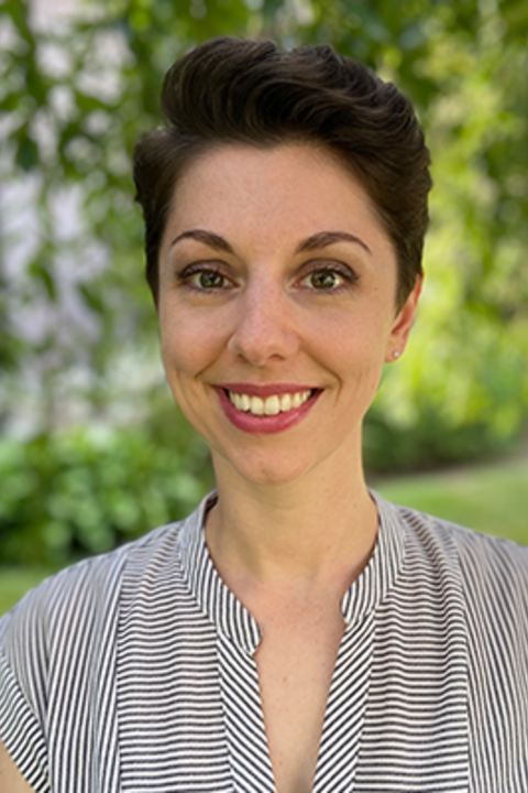A woman with short hair and pink lipstick wears a striped shirt and smiles directly at the camera. 