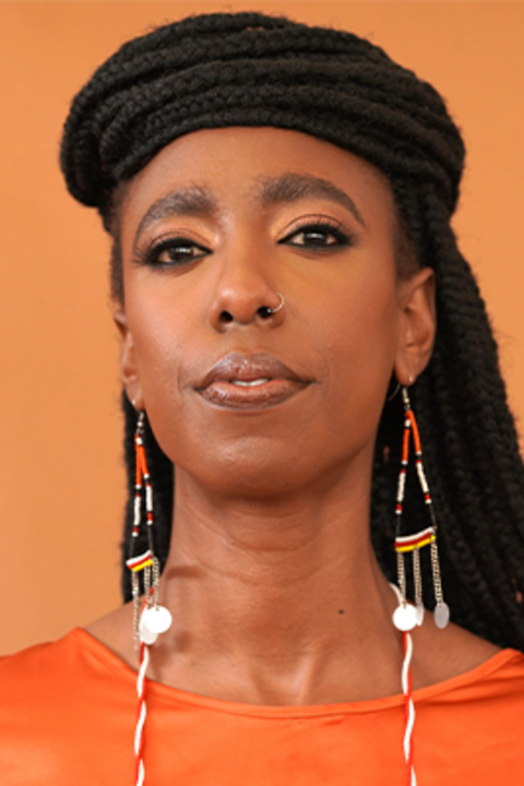 A woman with long black braids fall behind her head and are wrapped at the top of her head. She wears orange and has long beaded earrings.