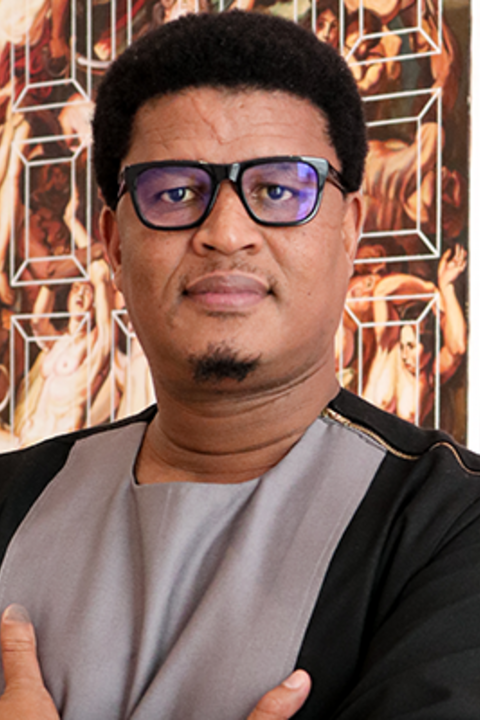 A man with dark skin and dark hair looks intensely with black glasses and a grey and black shirt.
