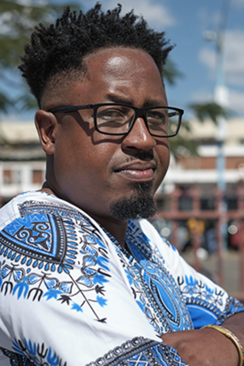 A man with dark skin and dark hair looks intensely over his shoulder with black glasses and a white and blue patterned shirt.