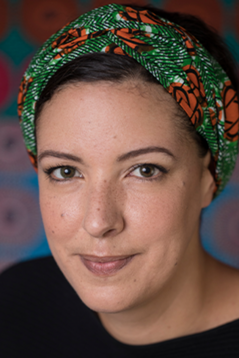 A woman with dark hair wears a colorful headband and black shirt while shirt smiling directly at the camera.