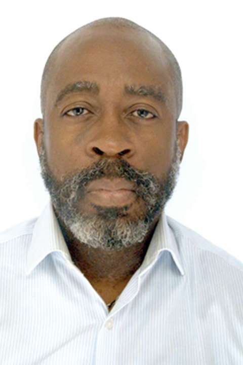 A bald man with dark skin and salt and pepper beard looks directly at the camera, wearing a white collared shirt.