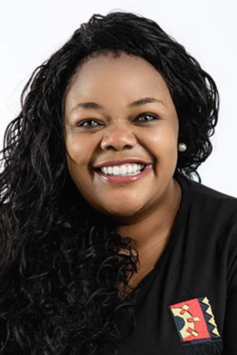 A woman in a black shirt with colorful pocket and long braids smiles at the camera.