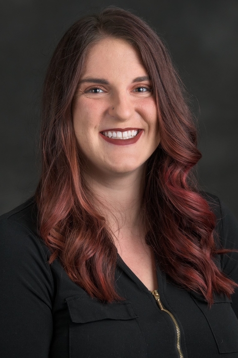 A woman with dark brown and red hair smiles at the camera in front of a dark background. 
