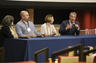 A panel of four people seated at a table with a red tablecloth. The individual on the far right speaks into a microphone. 