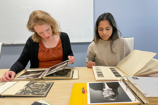 Two people sit at a table looking down at paper samples and books.