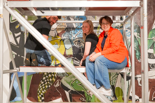 Three individuals sit on a scaffold in front of a colorful painting smiling at the camera.