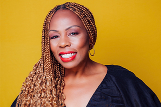 A woman with long braids and red lipstick smiles at the camera in front of a golden yellow background.