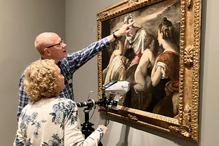 Two people look at a painting with a big gold frame while pointing and maneuvering a scientific instrument.