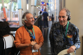 Two men in discussion, one wearing an orange shirt and the other in a blue patterned shirt and headphones.