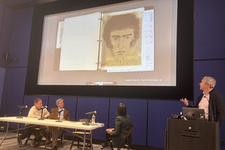 Panelists sit at a table looking at a projection while a speaker presents from a podium.