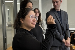 A woman in all black and glasses holds up and examines a small sample.