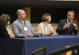 Four panelists seated at a blue table engaged in discussion. One speaking into a microphone.