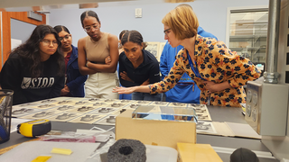 Individuals leaning over a spread of photographs that are varying in paper color. 