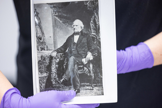 Black and white photograph of a seated man held by someone wearing purple gloves.