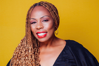 A woman with long braids and red lipstick smiles at the camera in front of a golden yellow background.
