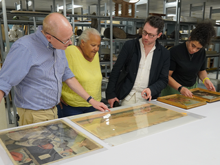 Four people look at artworks laying on a table in a storage facility.