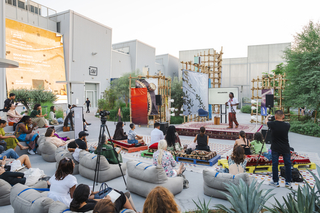 A group sits in an outdoor seating area facing a stage with a presenter. 