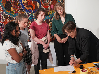 A woman in a black shirt explains a lesson to a group of students looking down at a table.