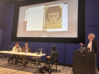 Panelists sit at a table looking at a projection while a speaker presents from a podium.