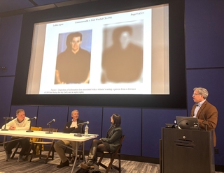 A man stands behind a podium during a panel discussion with wo images of men on the screen.